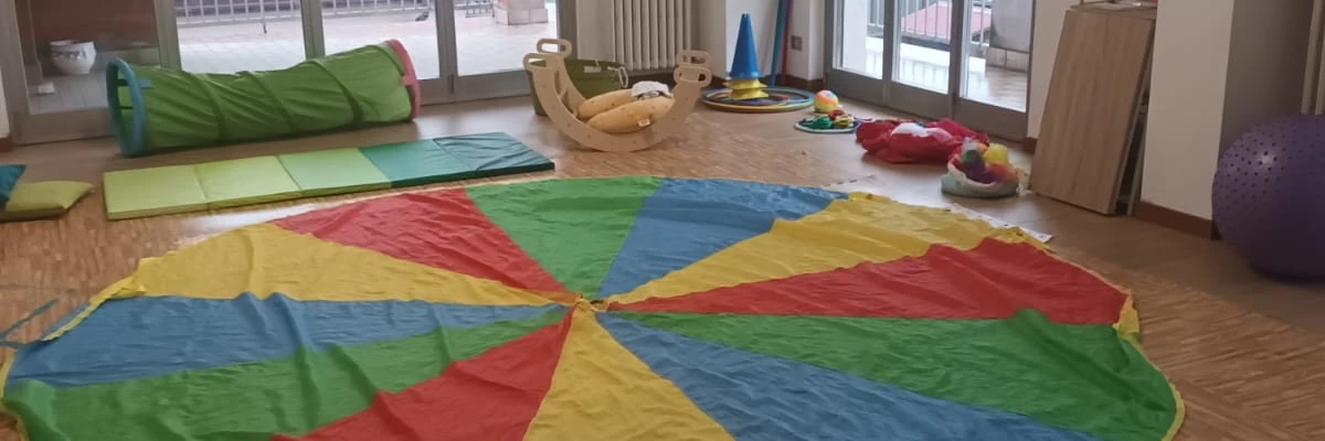 tappeto rotondo di tanti colori steso nella sala di psicomotricità, con i vari materiali intorno come cerchi, coni, stoffe, palla, cuscini, tunnel