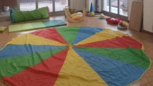 tappeto rotondo di tanti colori steso nella sala di psicomotricità, con i vari materiali intorno come cerchi, coni, stoffe, palla, cuscini, tunne
